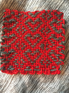 A red woven coaster, featuring gray outlined hearts and diamond motifs on it, front view