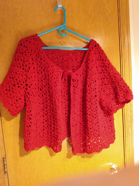 A red crocheted lacy cropped cardigan with an open front and a tie closure at neck.