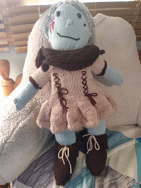 A knitted doll, front view. She is wearing a pleated brown tunic, knitted boots, and a knitted cape